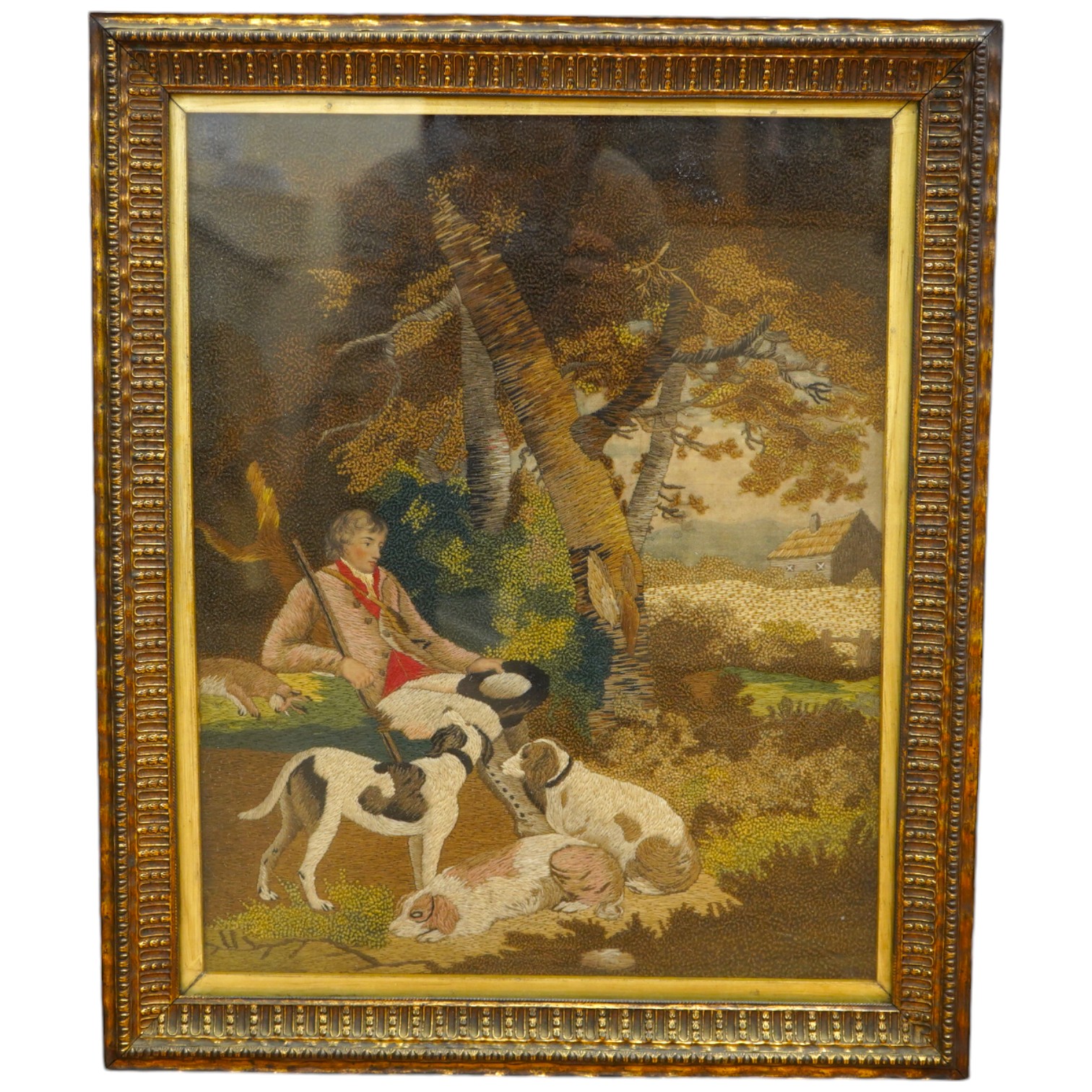 A framed early 19th century wool worked embroidery of huntsman sitting with a rabbit and his three hunting dogs under a tree, by a stream, with a field and cottage in the background, the embroidery worked in fine multi-c